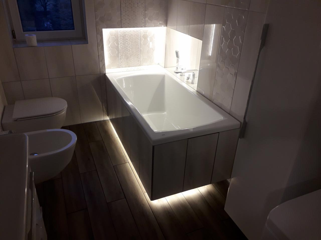 Nowoczesna łazienka z podświetlaną wanną zabudowaną drewnopodobną okładziną, mozaiką na ścianie i drewnianą podłogą, oświetlenie LED podkreśla krawędzie wanny i wnęki.