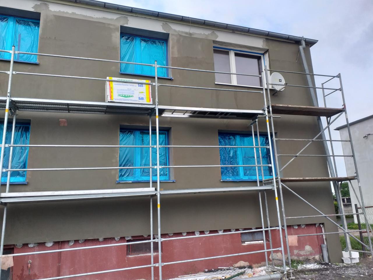 Dwupiętrowy budynek w trakcie prac elewacyjnych; okna zabezpieczone niebieską folią, rusztowanie, świeżo nałożony szary tynk, widoczna antena satelitarna.