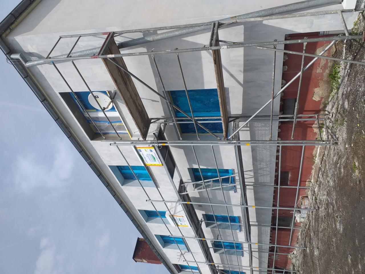 Budynek w trakcie ocieplania elewacji z widocznymi rusztowaniami, zabezpieczonymi oknami folią i fragmentem elewacji w kolorze ceglastym.