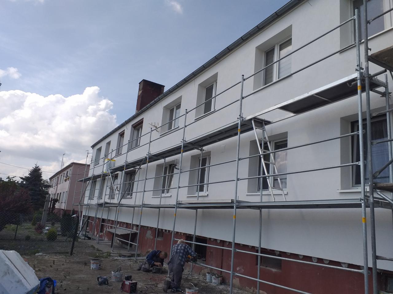 Budynek wielorodzinny w trakcie prac elewacyjnych, widoczne rusztowania, pracownicy przy gruncie, fragment otynkowanej ściany na biało i fragment ściany z widoczną izolacją termiczną...