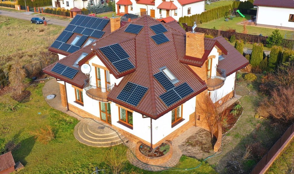 Widok z lotu ptaka na dom jednorodzinny z brązowym dachem, na którym zamontowane są panele słoneczne; widoczna antena satelitarna, zielony trawnik i sąsiednie domy.