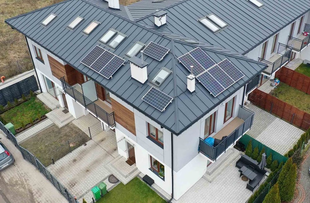 Widok z góry na nowoczesny dom dwurodzinny z szarą blachodachówką, na której zamontowano panele fotowoltaiczne wokół okien dachowych i kominów.