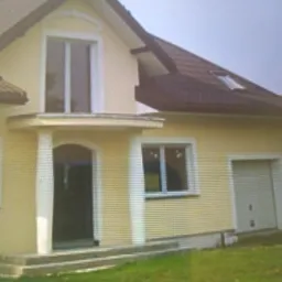 Żółty dom jednorodzinny z brązowym dachem, widoczne okna z białymi ramami, drzwi wejściowe z kolumnami i garaż z bramą segmentową.