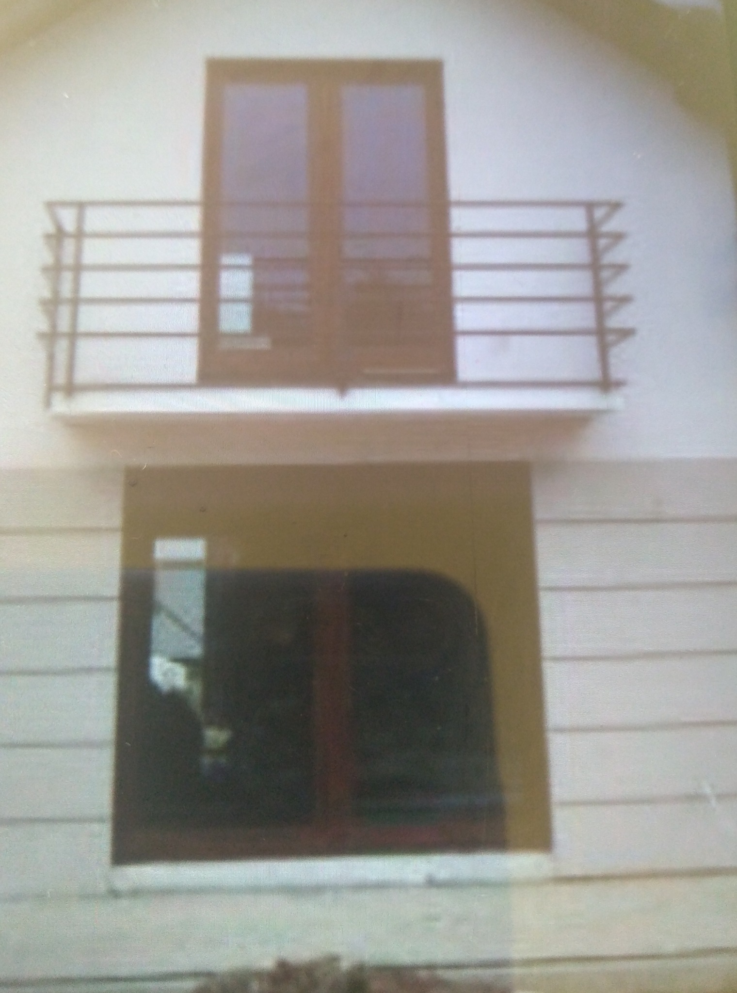 Dwukondygnacyjny fragment elewacji budynku z balkonem z brązową balustradą i oknem na parterze, z widoczną strukturą desek elewacyjnych.