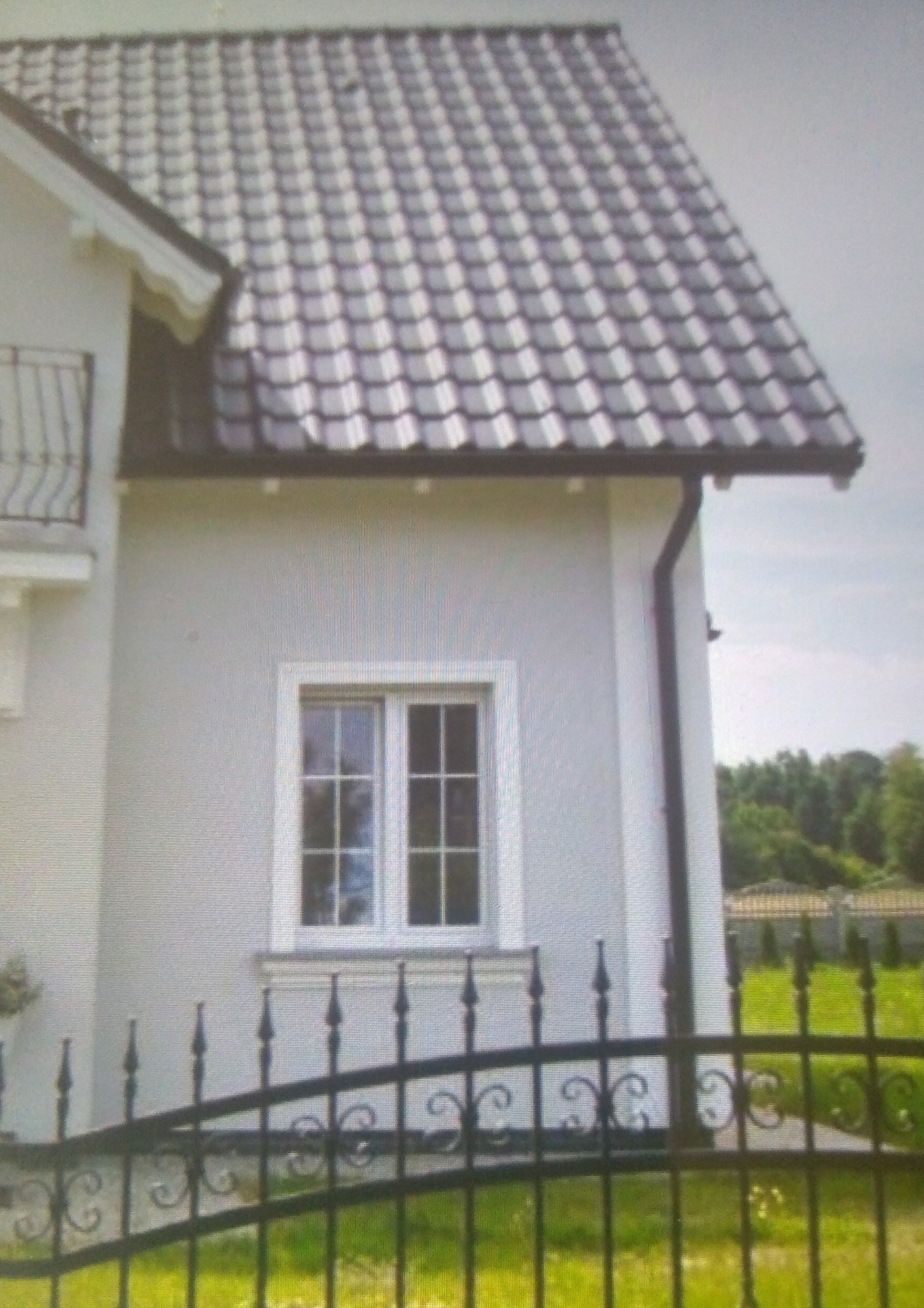 Fragment jasnej elewacji domu z oknem i ciemną dachówką, widoczny ozdobny metalowy płot