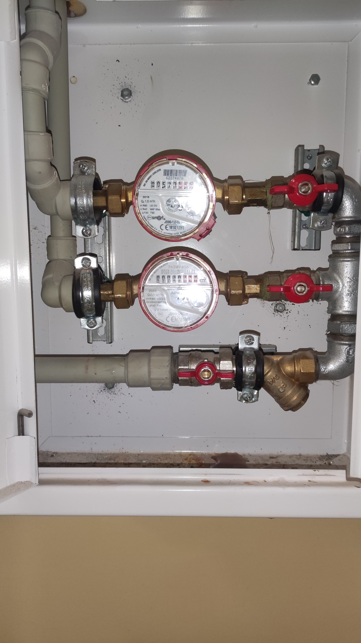 Trzy liczniki wody zamontowane w białej szafce, z widocznymi rurami z tworzywa i mosiądzu, zaworami z czerwonymi pokrętłami oraz elementami mocującymi.