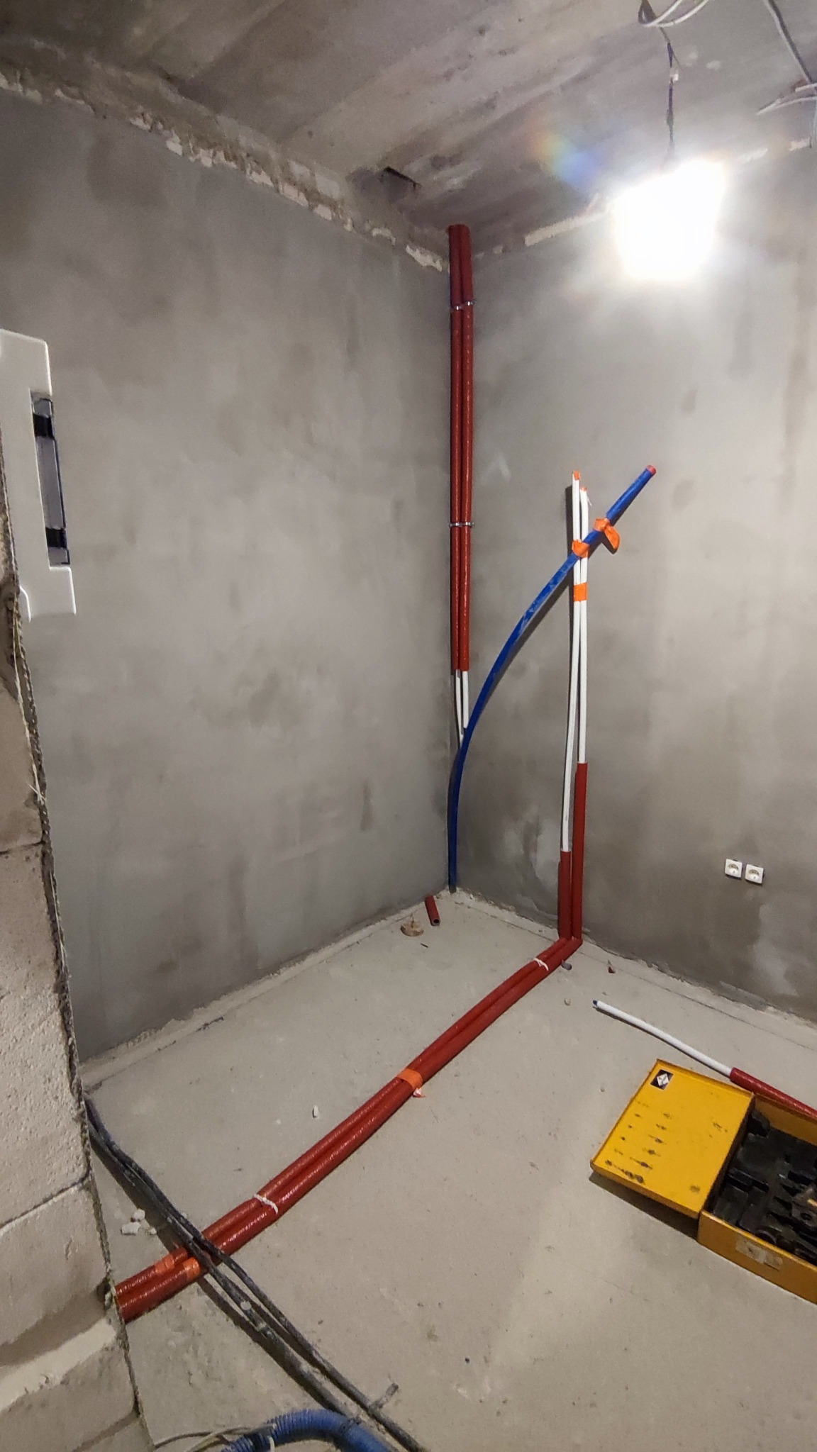 Surowy stan instalacji w nowym budynku: pionowe i poziome rury w izolacji termicznej, niebieska rura giętka, żółta skrzynka z narzędziami na szarym, betonowym podłożu.