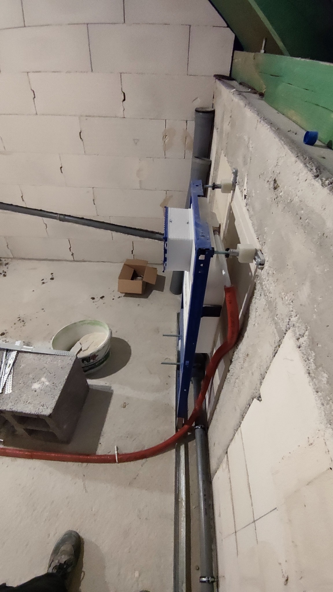 Stelaż podtynkowy WC zamontowany przy ścianie z betonu komórkowego, widoczne rury doprowadzające wodę, kanalizacja oraz elementy mocujące, w tle wiadro z zaprawą i karton.