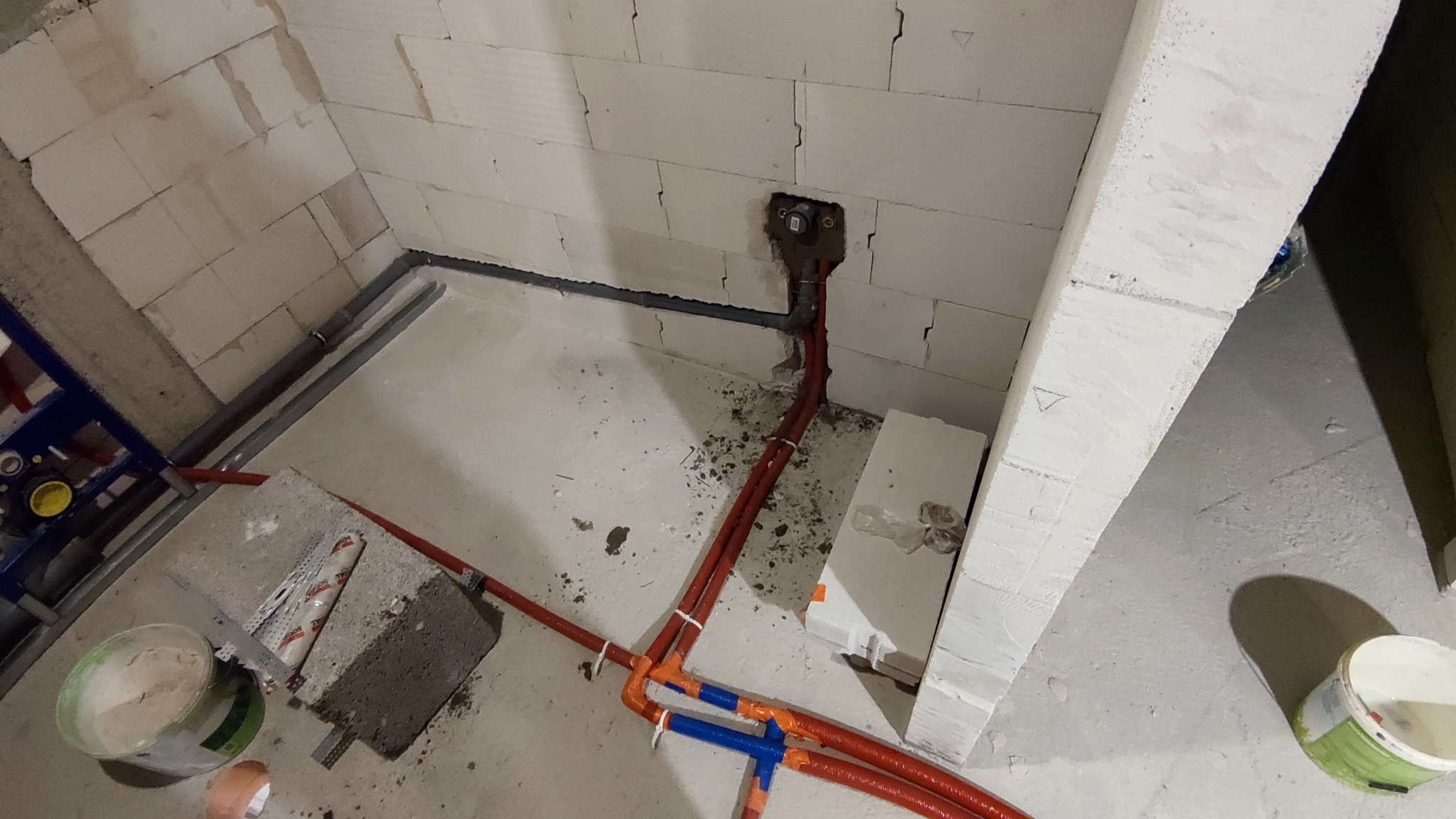 Surowy stan instalacji hydraulicznej w nowym budynku, widoczne rury w kolorze czerwonym i niebieskim, mocowane do ściany z betonu komórkowego, z fragmentami gruzu na podłodze.