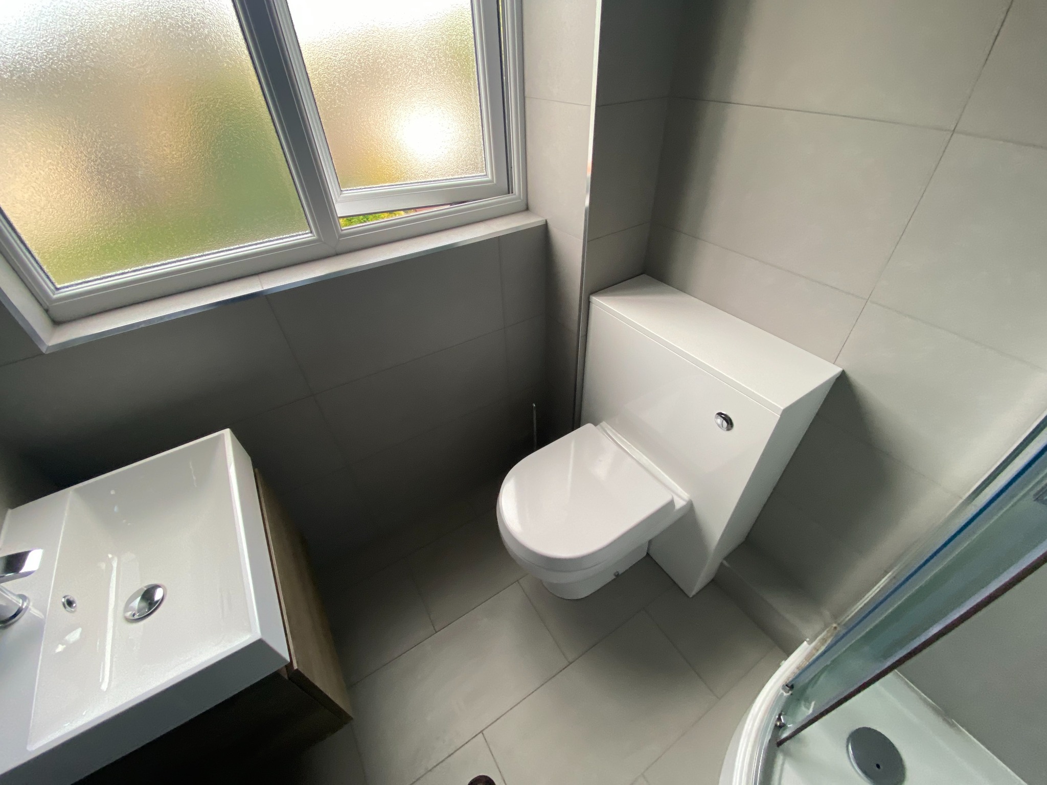 Nowoczesna łazienka w szarościach z białą ceramiką: umywalka nablatowa z szafką, kompakt WC i fragment kabiny prysznicowej. Okno z matową szybą w tle.