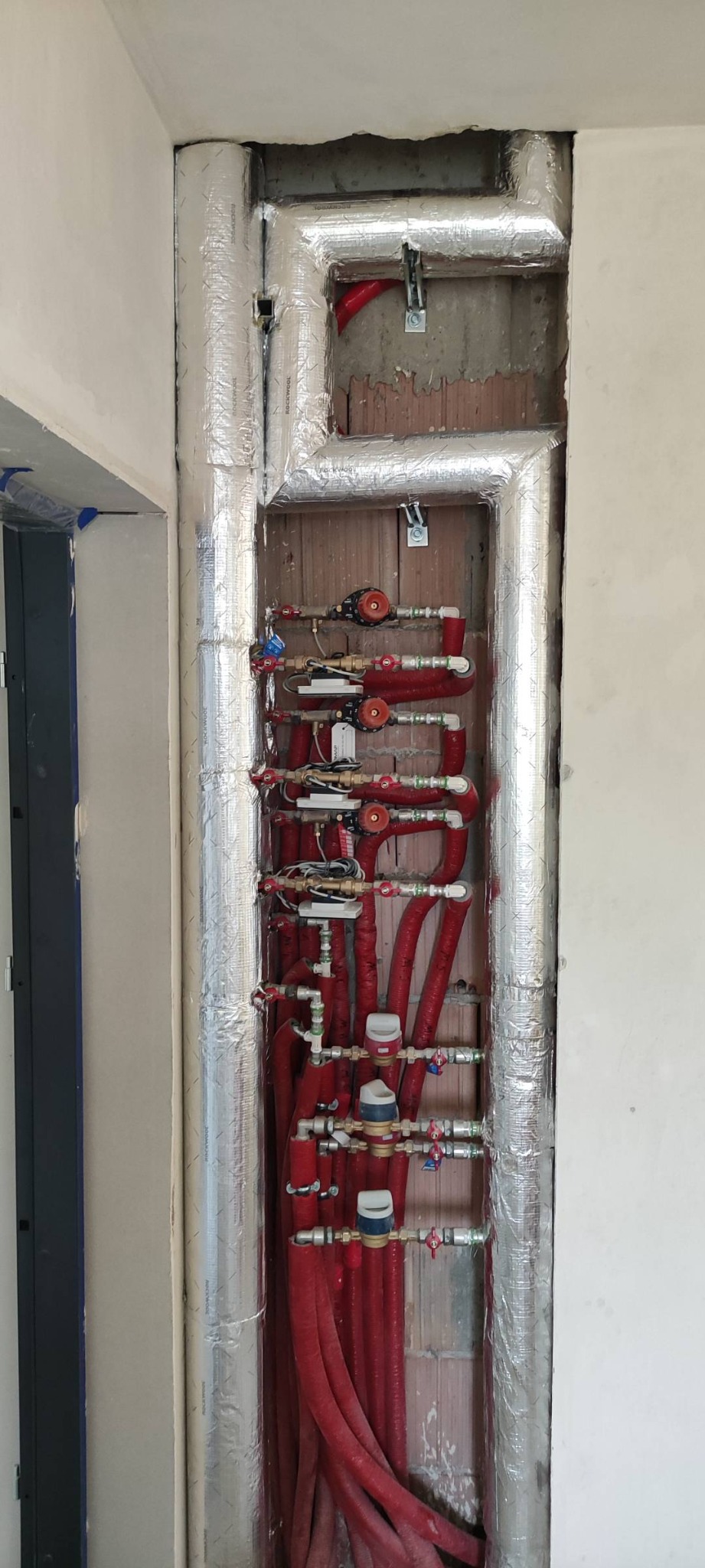 Czerwone rury centralnego ogrzewania z zaworami i licznikami, częściowo schowane w ścianie i zaizolowane srebrną folią, widok w trakcie montażu.