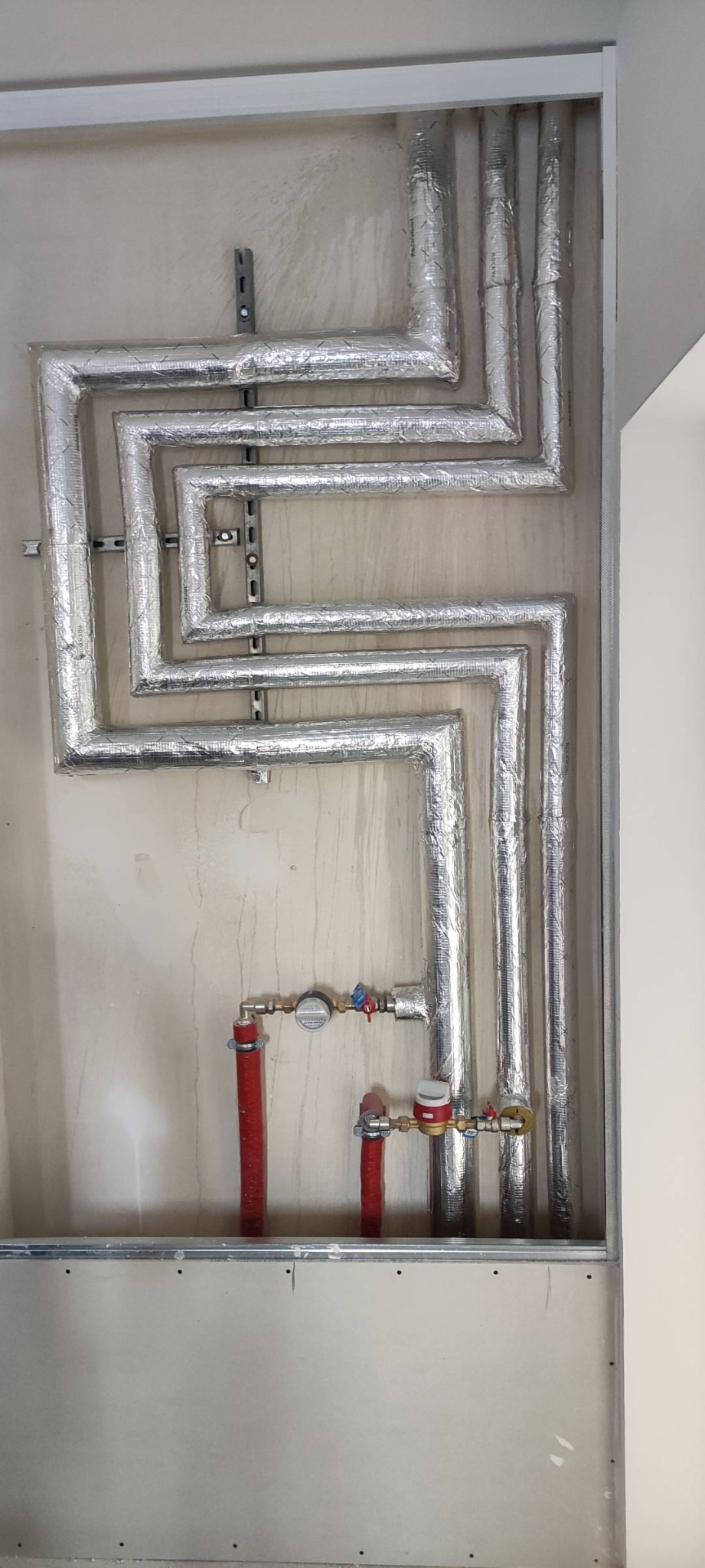 Instalacja rur grzewczych z izolacją termiczną i licznikami w Siedliszowicach, widoczne mocowania do ściany i fragment surowej płyty kartonowo-gipsowej.