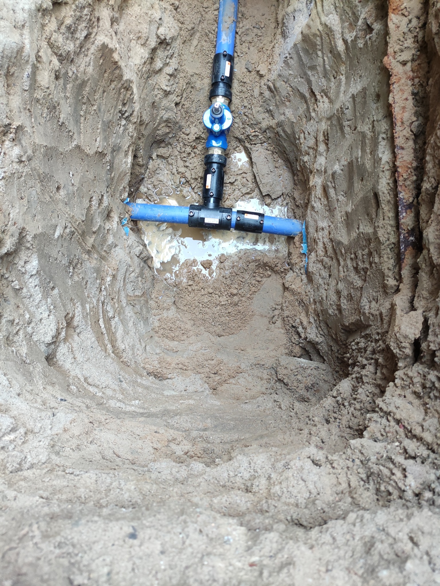 Niebieskie rury wodociągowe z czarnymi złączkami w wykopie ziemnym, widoczne ślady lekkiego zalania.