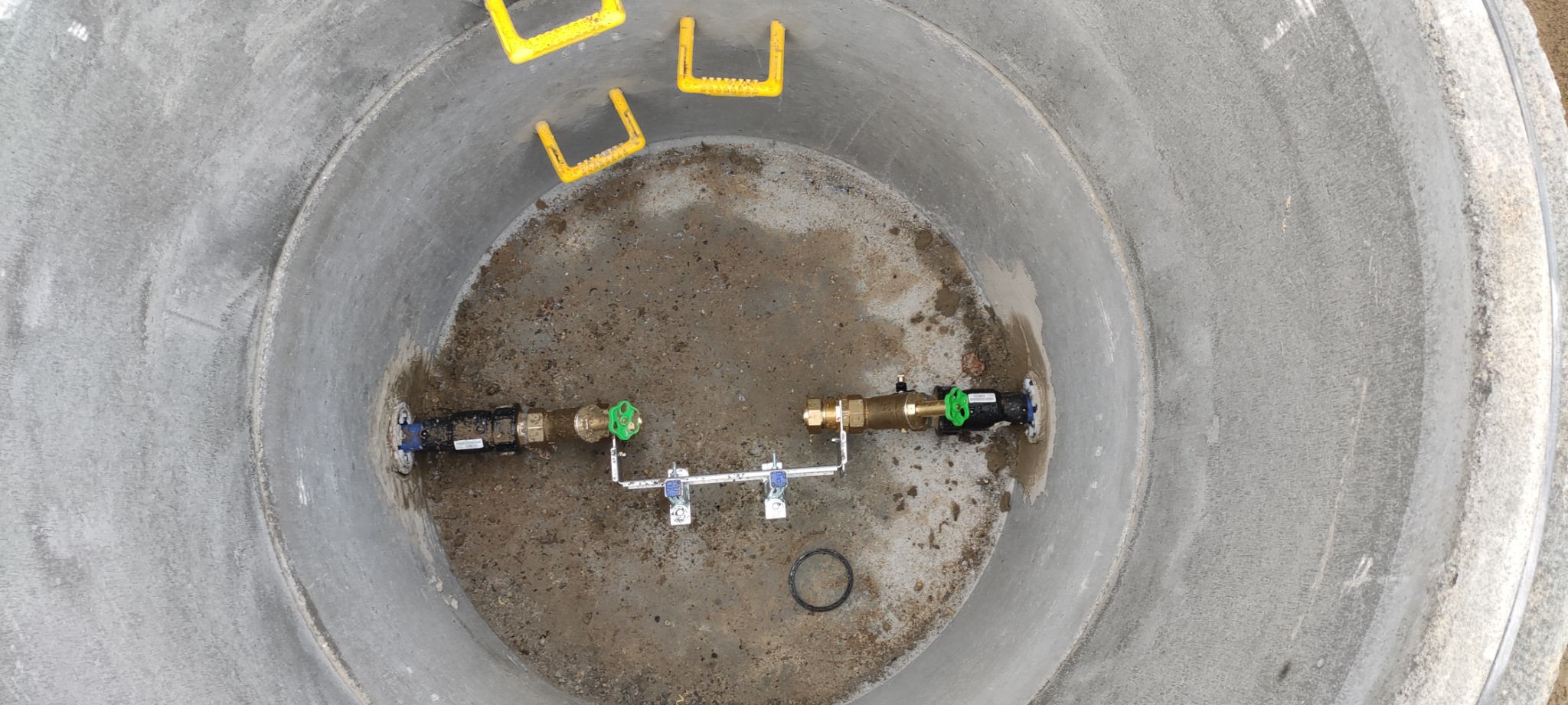 Wnętrze betonowej studni z zamontowanymi zaworami i rurami, widok z góry, żółte uchwyty w górnej części studni.