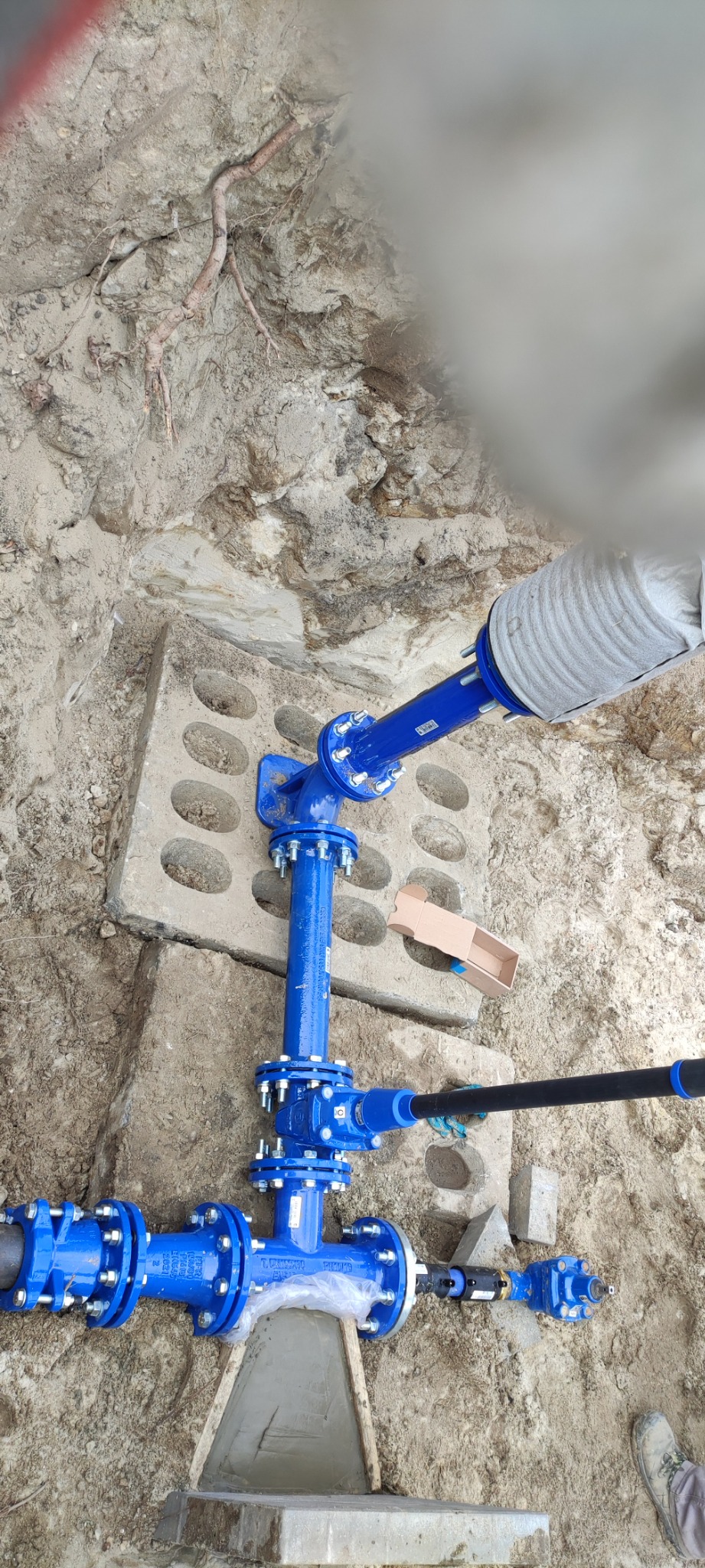 Niebieska instalacja wodociągowa z żeliwa sferoidalnego, częściowo zakopana w ziemi, z widocznymi złączkami kołnierzowymi i betonową podporą, zabezpieczona folią i kartonem.