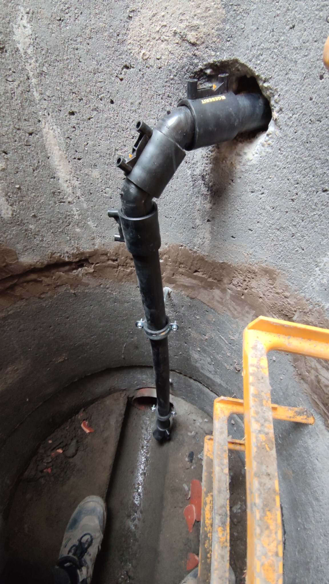 Czarna rura kanalizacyjna przechodząca przez otwór w betonowej studni, zabezpieczona obejmą, widoczny fragment drabiny i but pracownika.