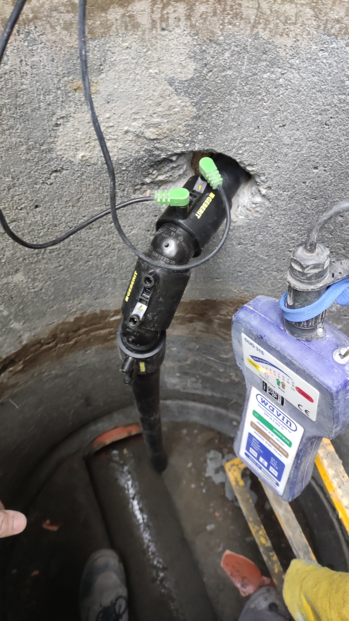 Czarny czujnik Geberit w otworze betonowej studni kanalizacyjnej, podłączony do urządzenia pomiarowego Wavin Duo 315 z żółtymi elementami drabiny w tle, wewnątrz widoczna rura.