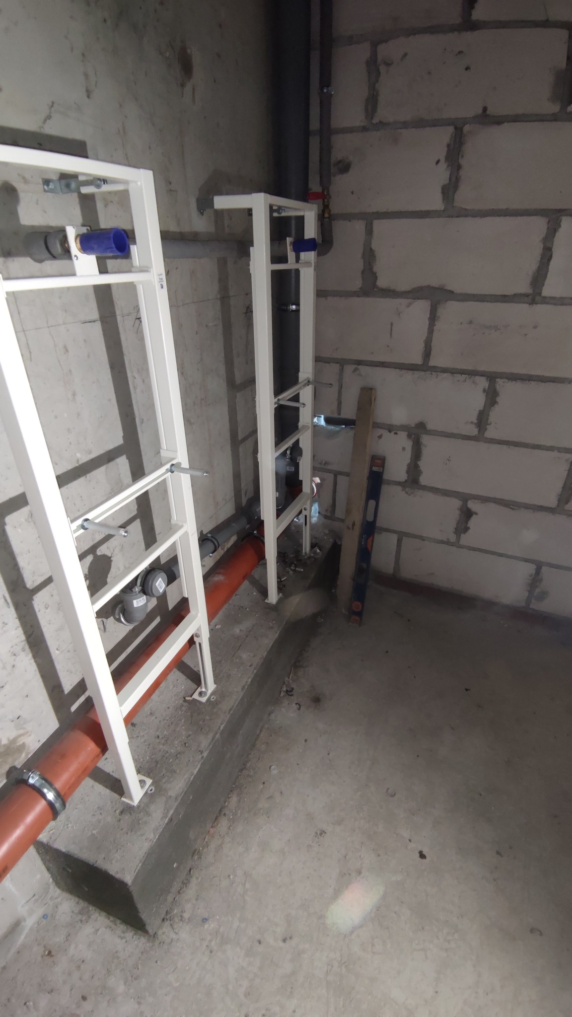 Dwa białe stelaże podtynkowe do WC z podłączonymi rurami kanalizacyjnymi z tworzywa, poziomica oparta o ścianę z betonu komórkowego.