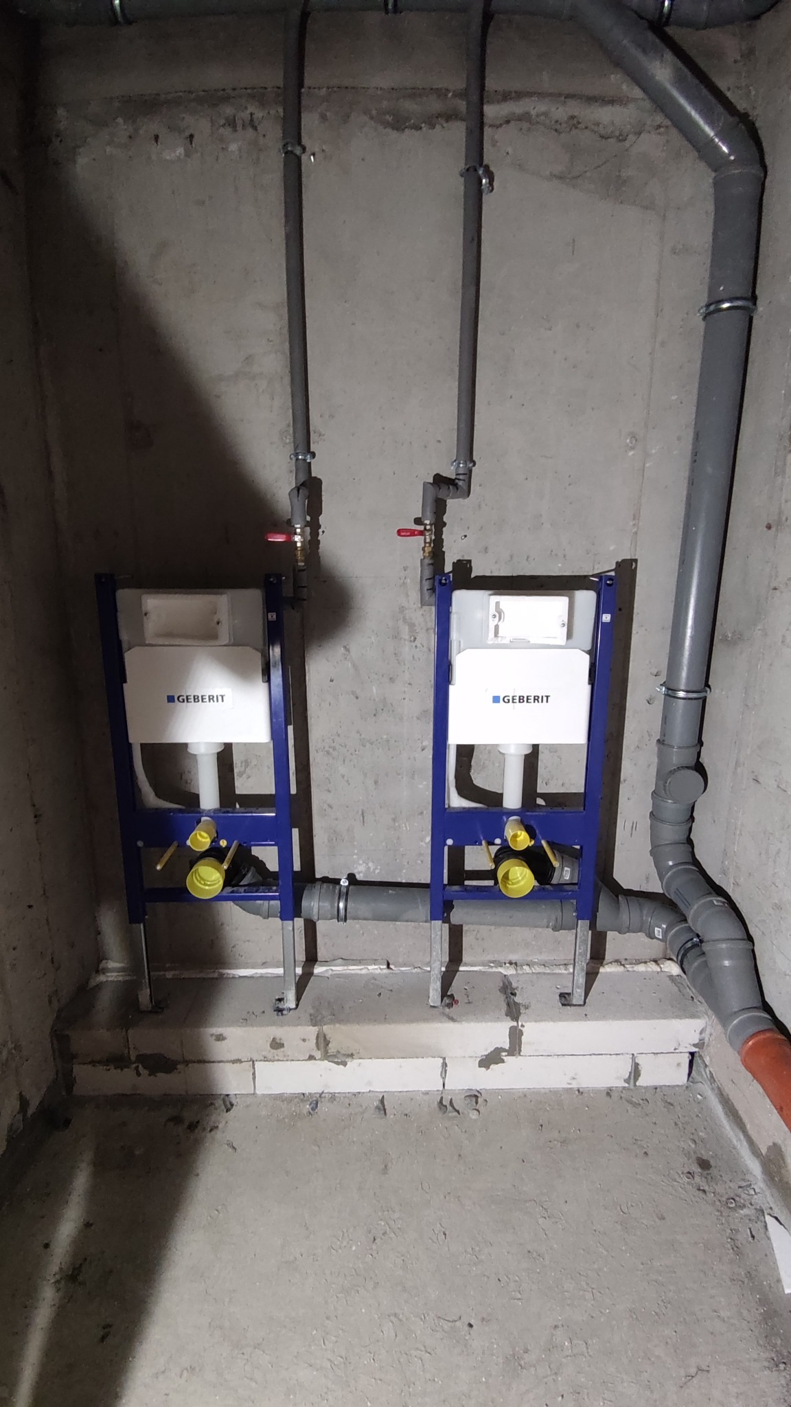 Dwa stelaże podtynkowe Geberit zamontowane w surowym pomieszczeniu, widoczne rury doprowadzające wodę i odpływ kanalizacyjny, ściany betonowe.