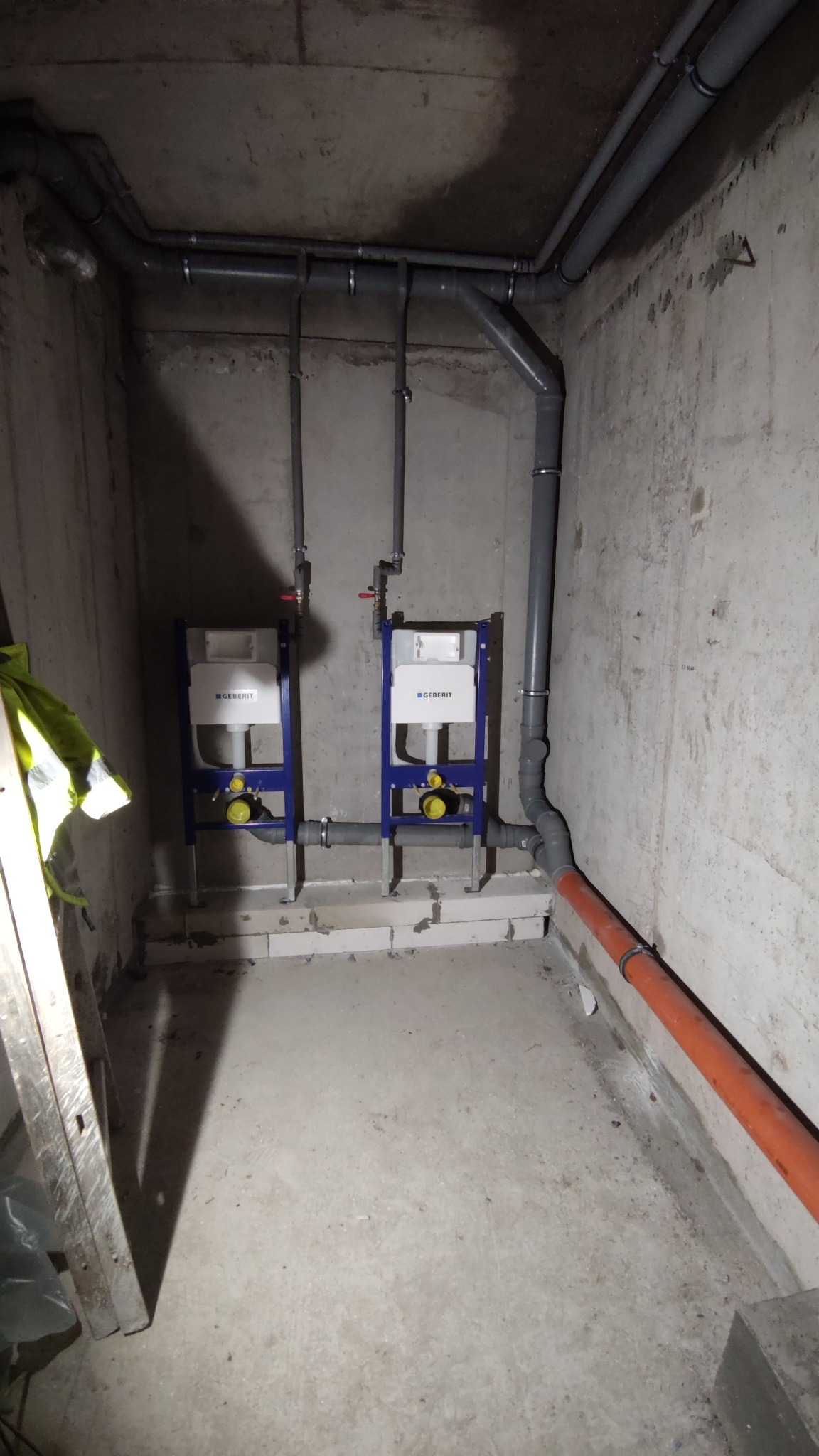 Dwa stelaże podtynkowe Geberit zamontowane na surowej ścianie z widocznymi szarymi rurami instalacji wodno-kanalizacyjnej i fragmentem drabiny z lewej strony.