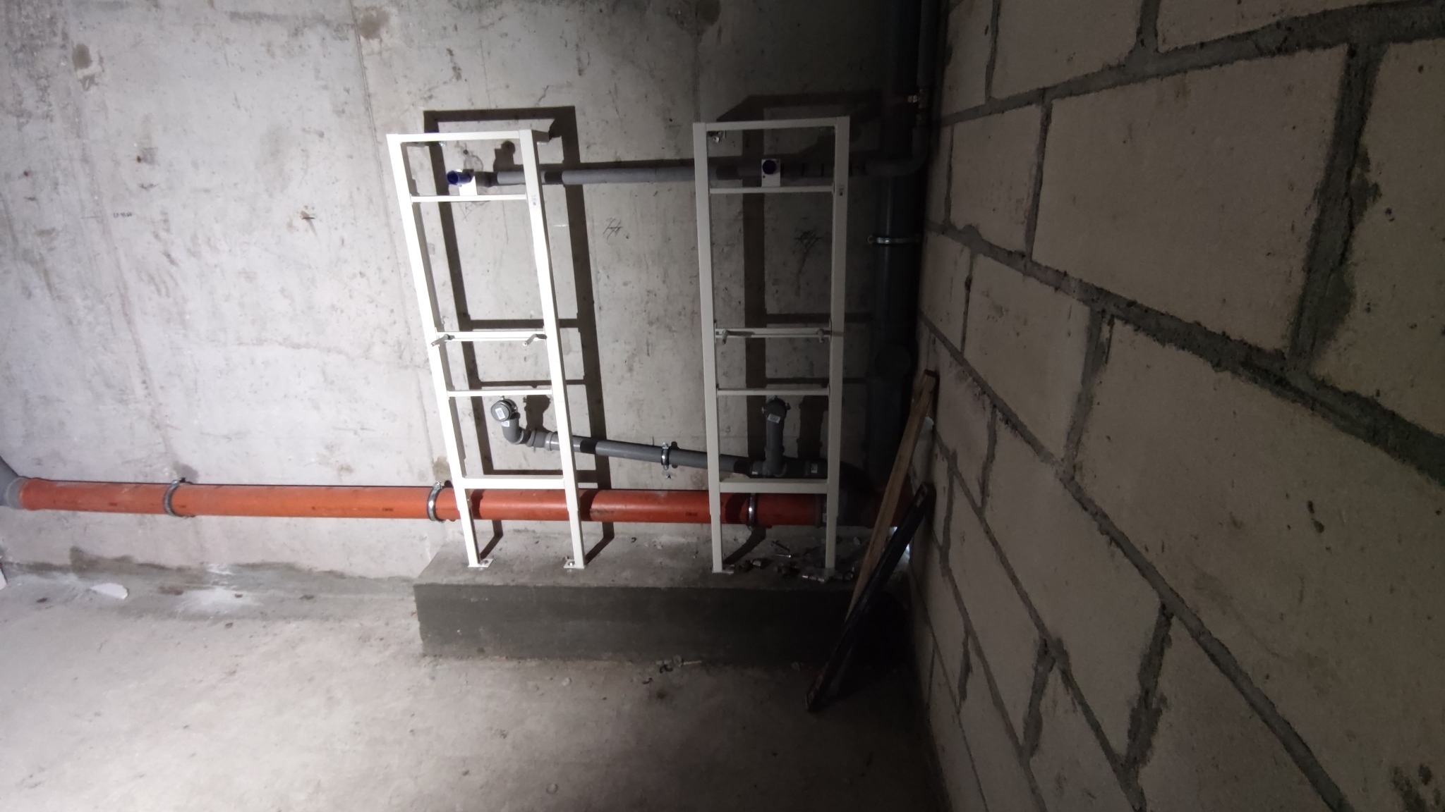 Surowy stan instalacji hydraulicznej w piwnicy: widoczne rury kanalizacyjne w kolorze terakoty i szare, zamontowane w metalowych stelażach przy betonowej ścianie z bloczków.