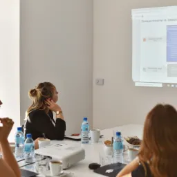 Sala konferencyjna z uczestnikami szkolenia, na ekranie wyświetlony slajd prezentacji dotyczący różnic między dostawą towarów a świadczeniem usług, na stole woda, kawa i przekąski.