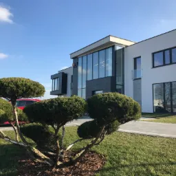 Nowoczesny budynek biurowy z przeszkloną fasadą i logo firmy, otoczony starannie przyciętymi krzewami i zielonym trawnikiem w słoneczny dzień.