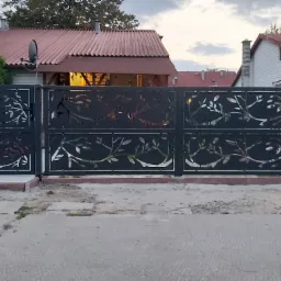 Czarna brama wjazdowa przesuwna z dekoracyjnym wycinanym laserowo motywem roślinnym na tle domu z czerwoną dachówką.