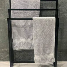 Czarny, minimalistyczny stojak na trzy szare ręczniki z paskami, na tle szarej ściany z płytek.