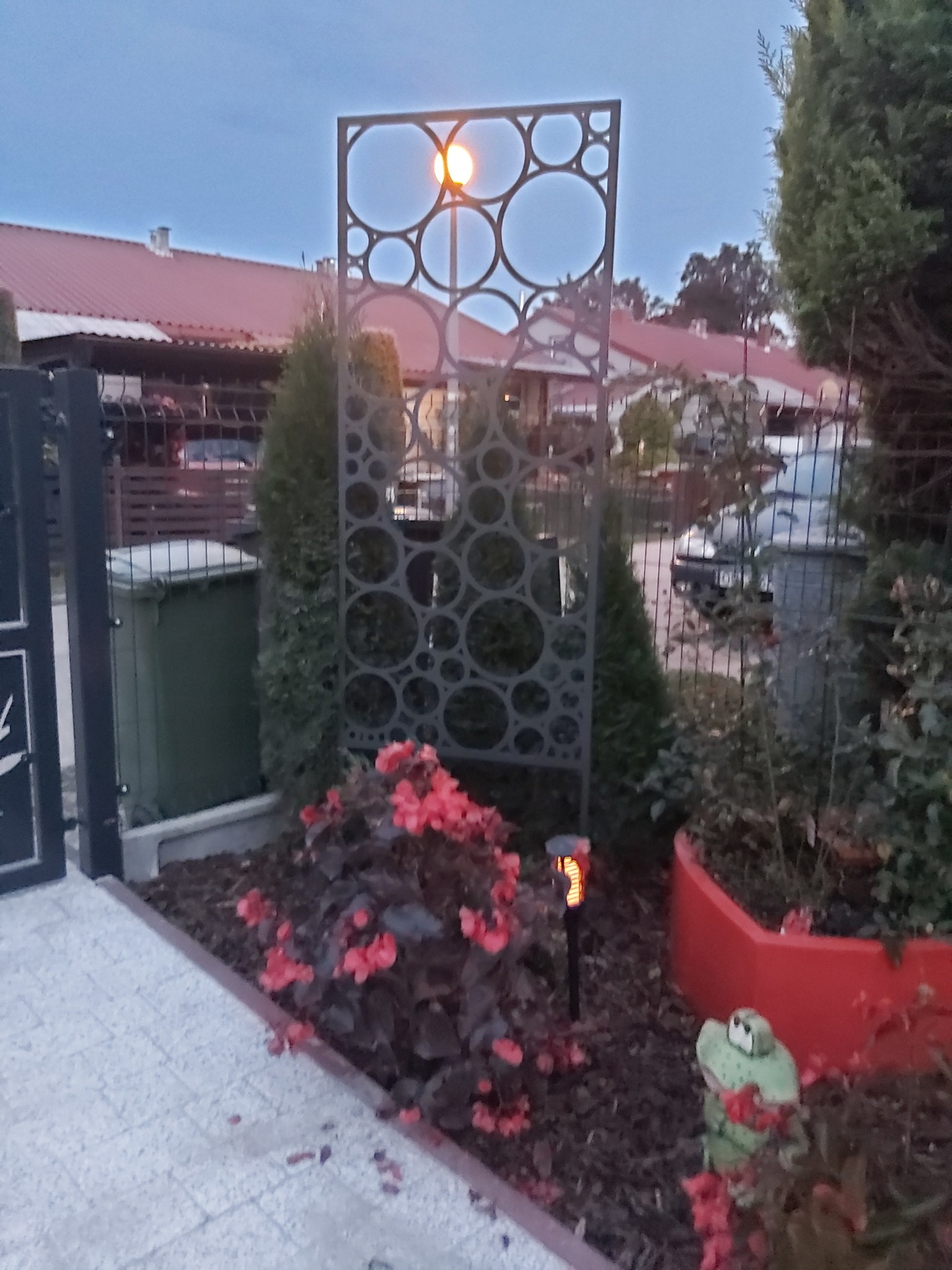 Dekoracyjny, metalowy panel z wyciętymi okręgami, oddzielający ogród od posesji, oświetlony lampą solarną, z widocznymi kwiatami i tujami w tle.