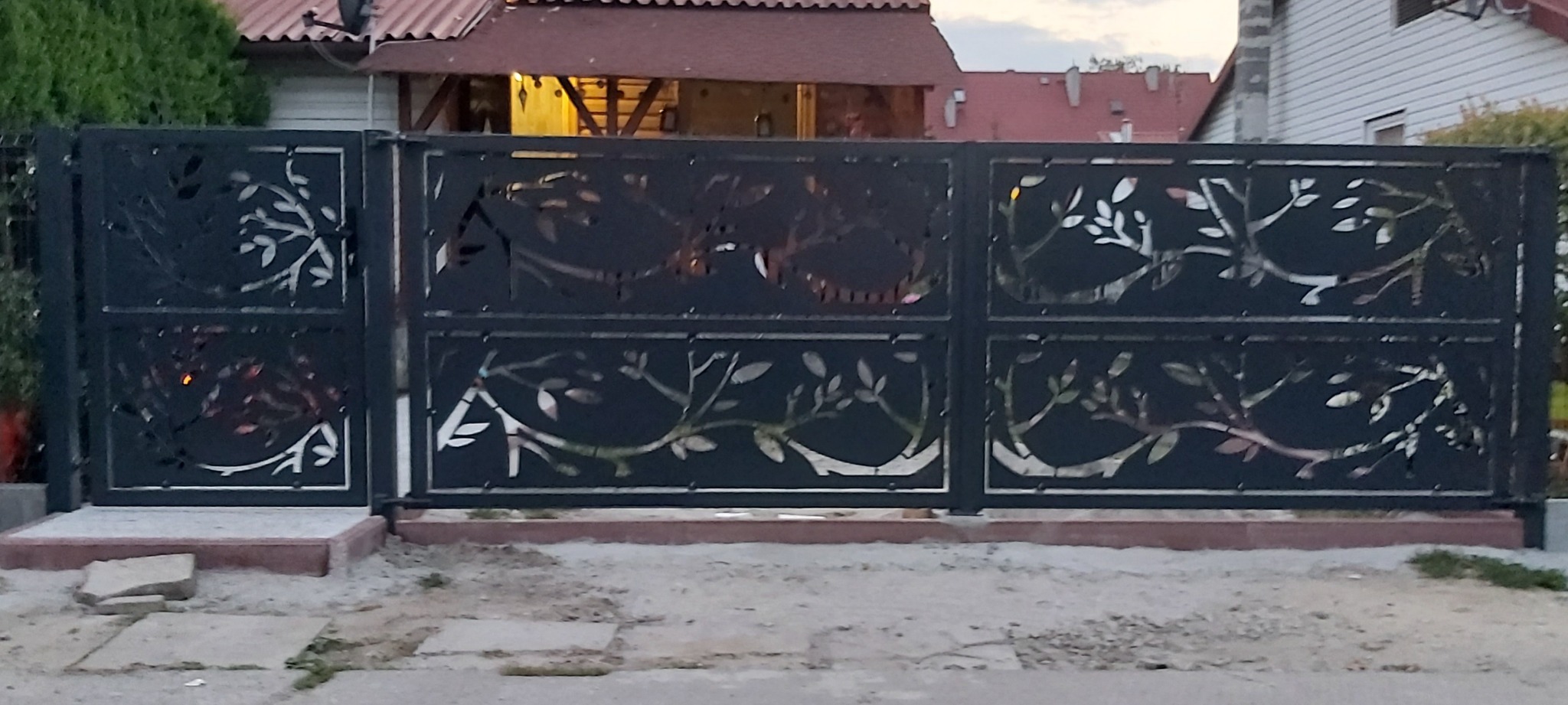 Czarna, metalowa brama wjazdowa z ażurowym motywem drzewa, osadzona na betonowej podstawie, przed domem z czerwonym dachem.