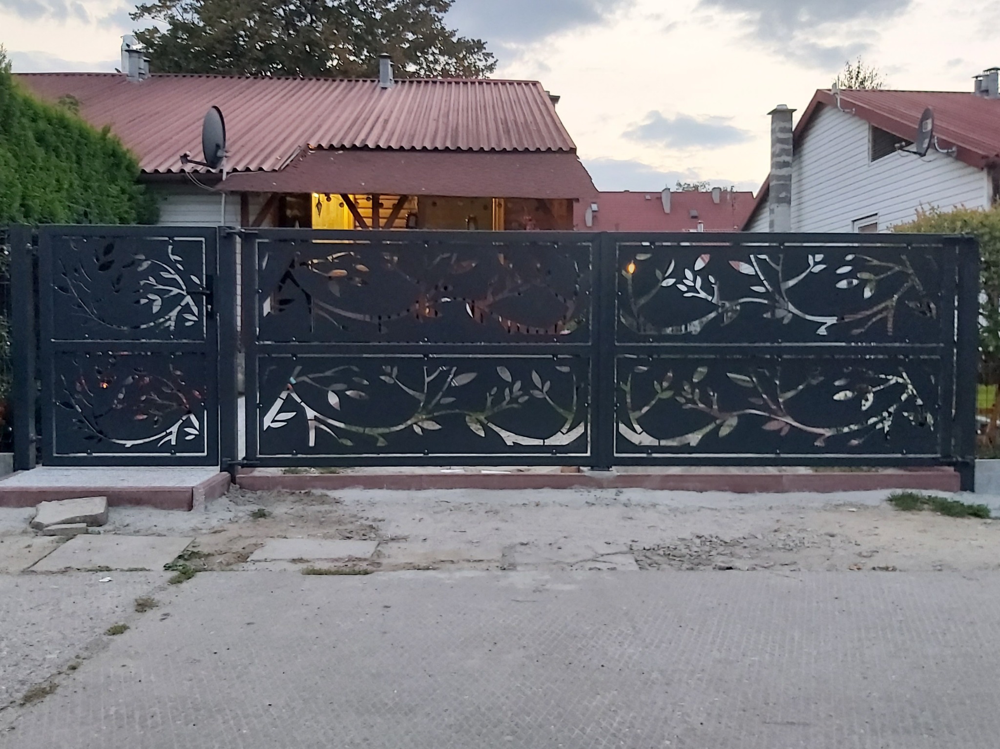Czarna brama wjazdowa przesuwna z dekoracyjnym wycinanym laserowo motywem roślinnym na tle domu z czerwoną dachówką.