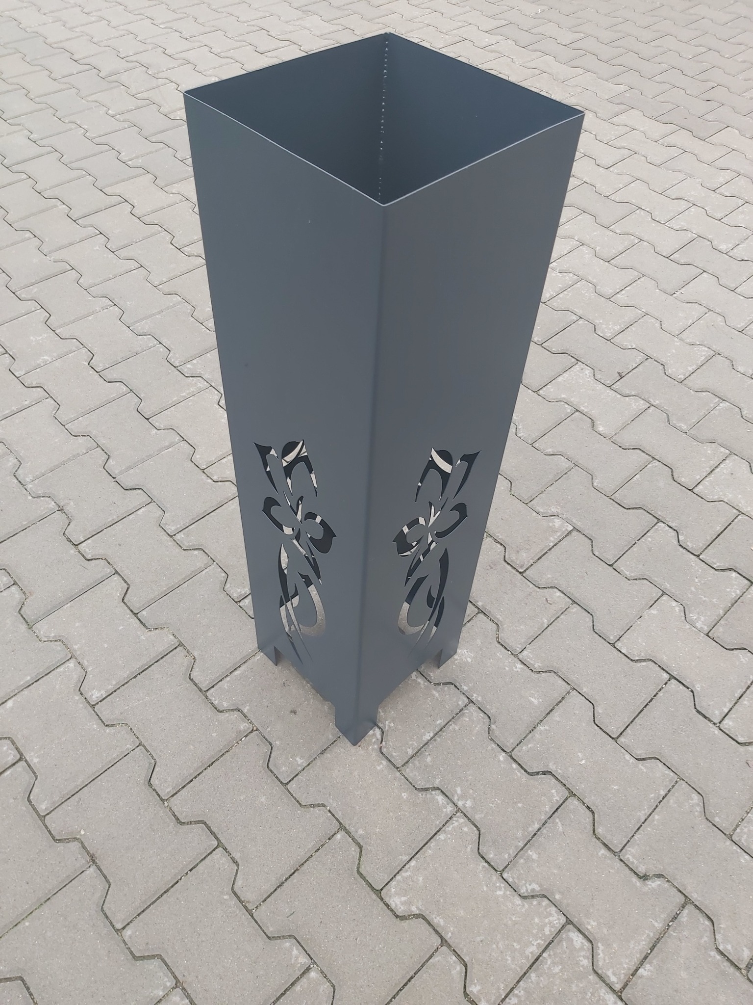 Szara, metalowa donica w kształcie prostopadłościanu z wyciętym laserowo, abstrakcyjnym wzorem kwiatowym, stojąca na szarej kostce brukowej.