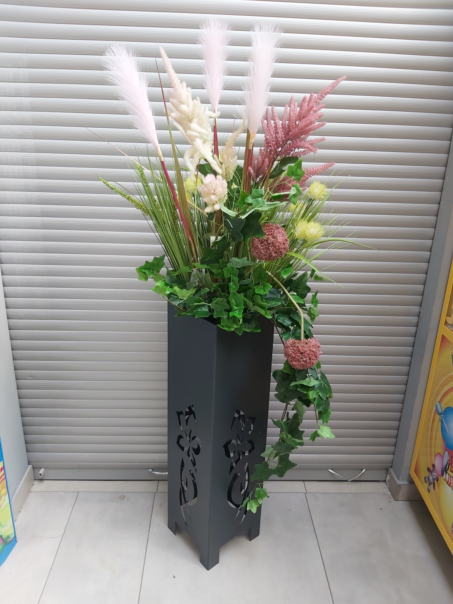 Dekoracyjna kompozycja kwiatowa w czarnym, ażurowym postumencie z motywem roślinnym, ustawiona na jasnej podłodze na tle szarej rolety.