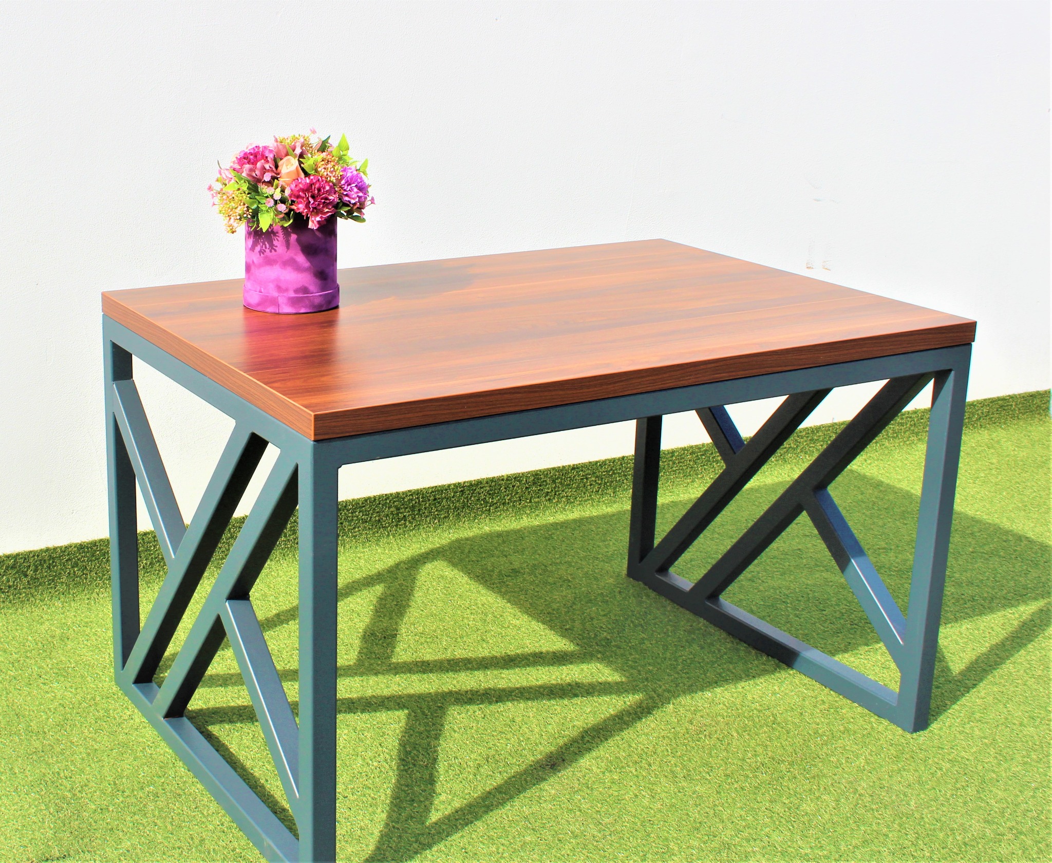Stół z drewnianym blatem i metalową podstawą w kształcie litery X, na sztucznej trawie, z fioletowym pudełkiem z kwiatami na blacie.
