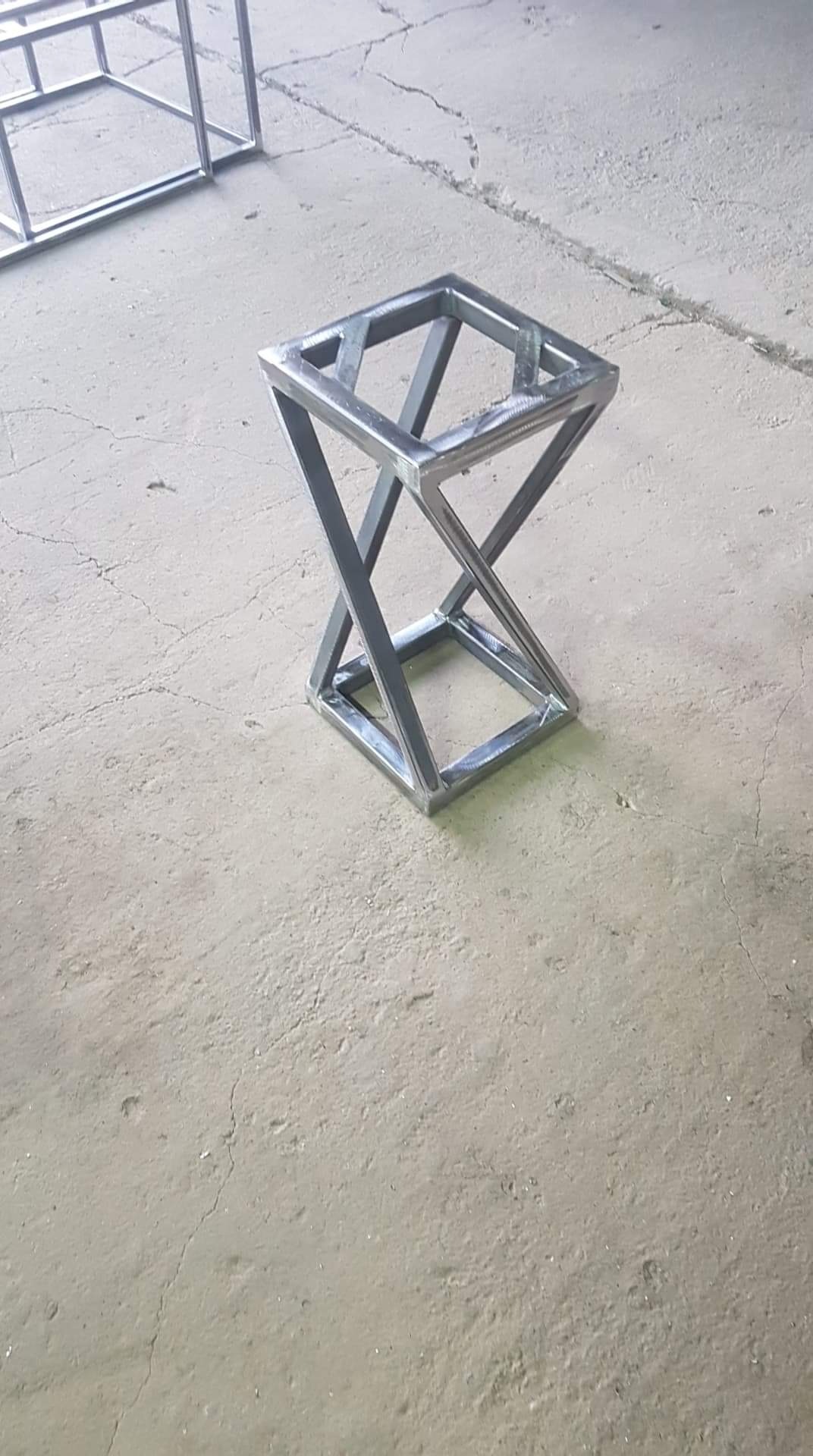 Metalowa konstrukcja spawana z profili kwadratowych, tworząca geometryczny kształt przypominający klepsydrę, surowe wykończenie, widoczne spawy, szary beton w tle.