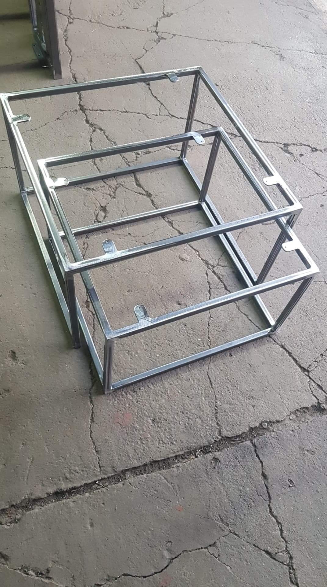 Dwa metalowe szkielety konstrukcyjne, prawdopodobnie do bram lub ogrodzeń, z widocznymi uchwytami montażowymi, leżące na betonowej posadzce z pęknięciami.