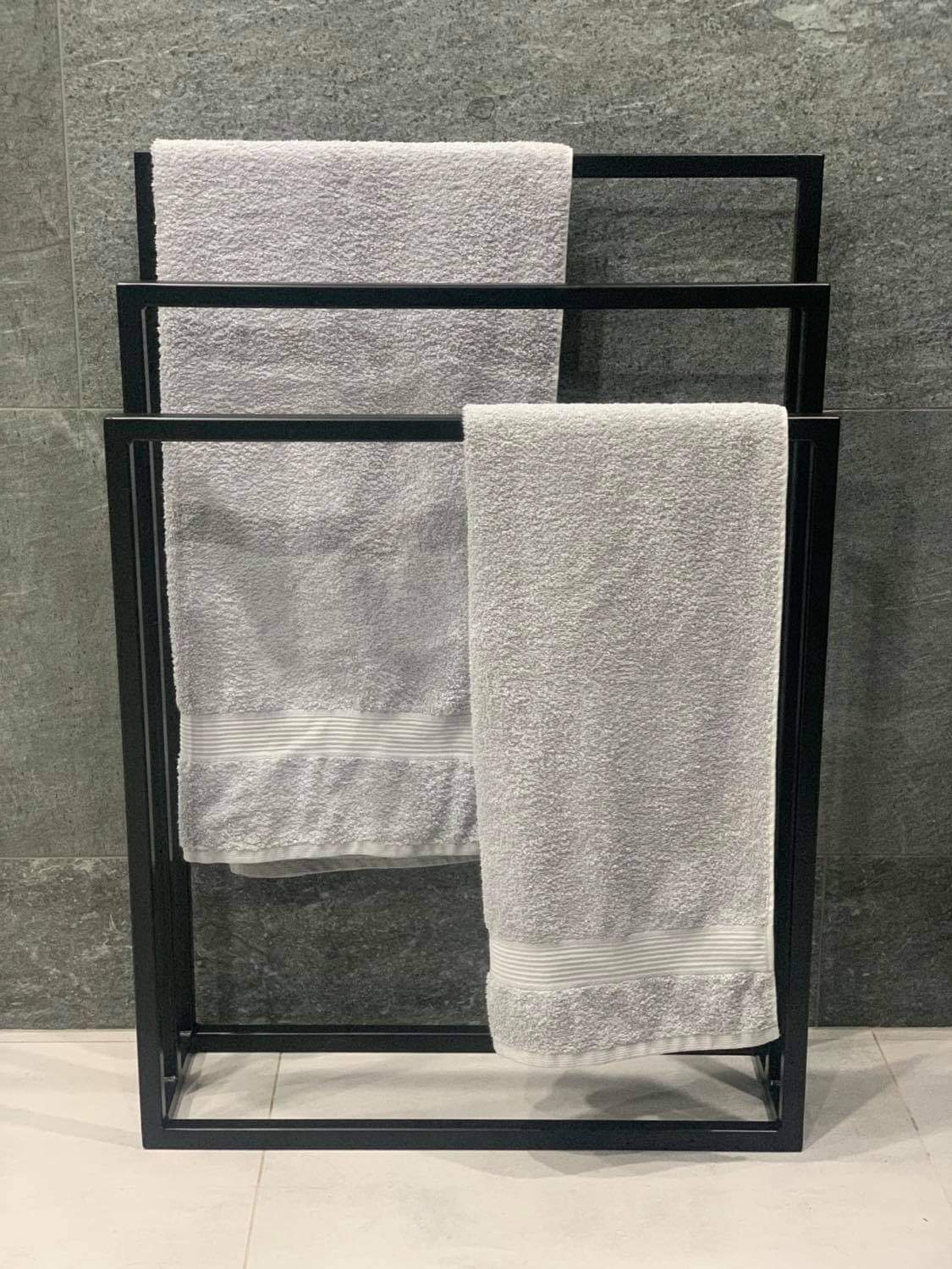 Czarny, minimalistyczny stojak na trzy szare ręczniki z paskami, na tle szarej ściany z płytek.
