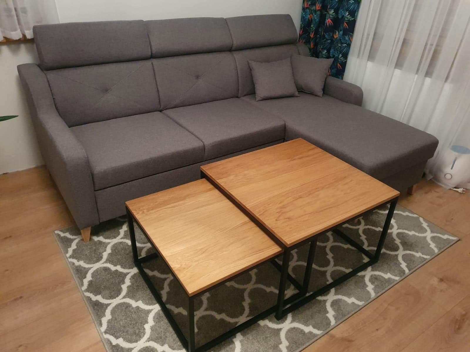 Szara sofa narożna z regulowanymi zagłówkami i dwoma drewnianymi stolikami kawowymi z czarnymi metalowymi nogami na szarym dywanie we wzory.