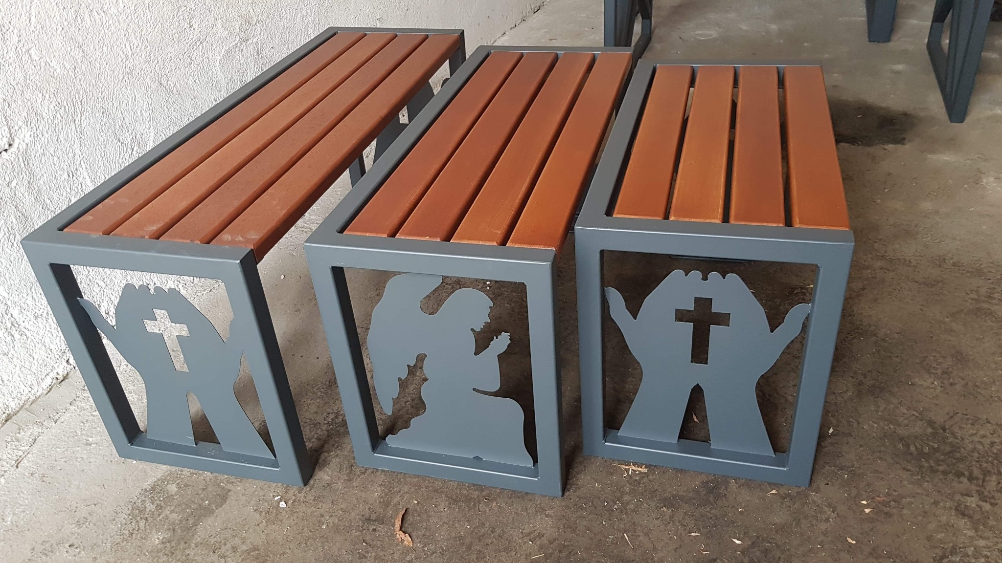 Trzy ławki z drewnianym siedziskiem i metalową ramą z wyciętymi motywami: dłonie z krzyżem i anioł w modlitwie.