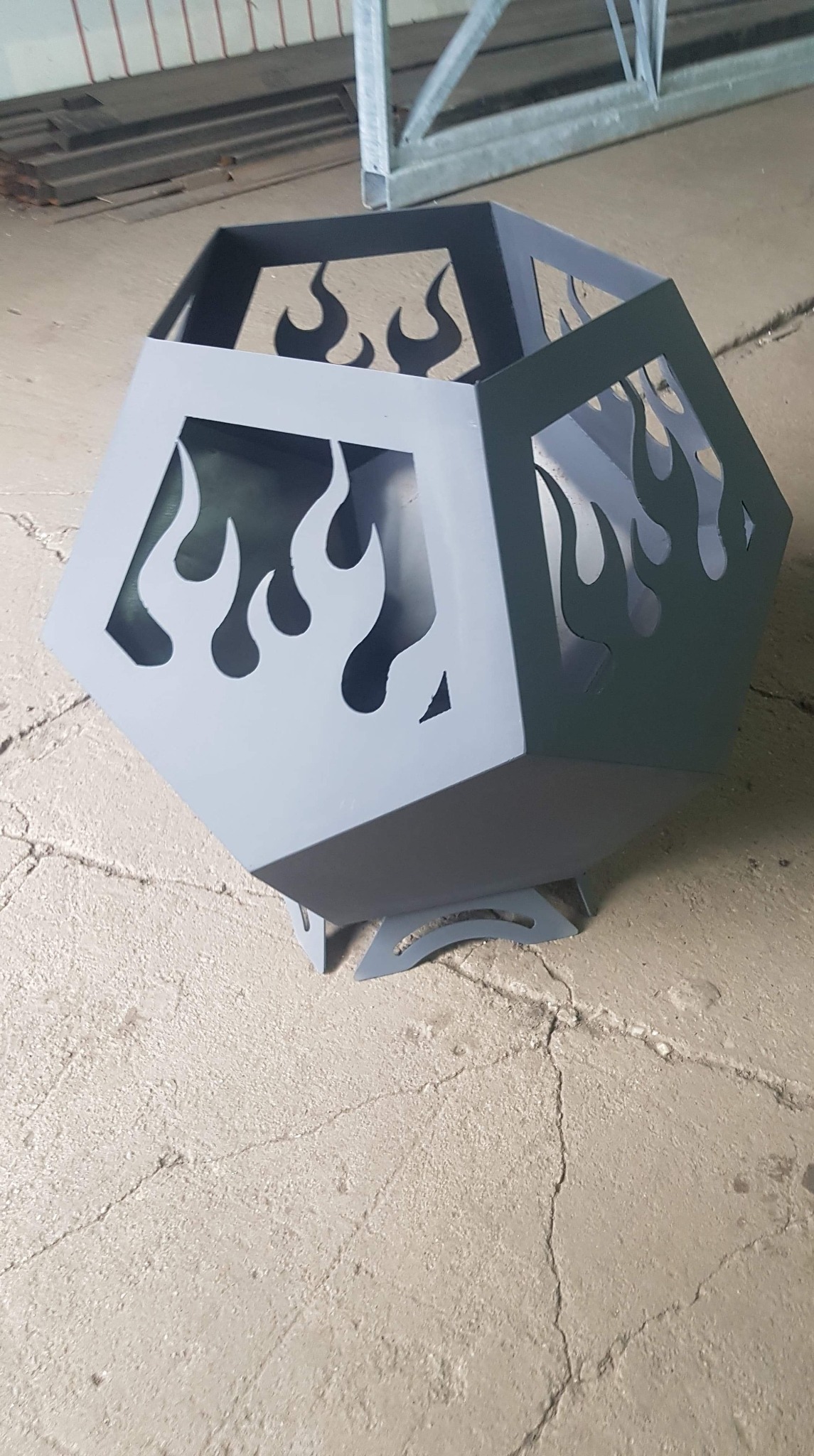 Szary, metalowy kosz na ognisko o heksagonalnym kształcie z wyciętymi motywami płomieni, stojący na trzech krótkich nóżkach, na betonowej posadzce.