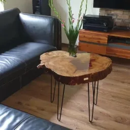 Stolik kawowy z blatem z naturalnego drewna z żywicą epoksydową, osadzony na trzech metalowych nogach typu hairpin, obok czarnej skórzanej sofy w salonie z drewnianą podłogą i szafką RTV z ceglaną...