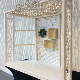 Drewniany bufet w stylu rustykalnym z napisem 'Bufet Wiejski', oświetlony girlandą żarówek, z dekoracjami w postaci wianka i rysunków warzyw.