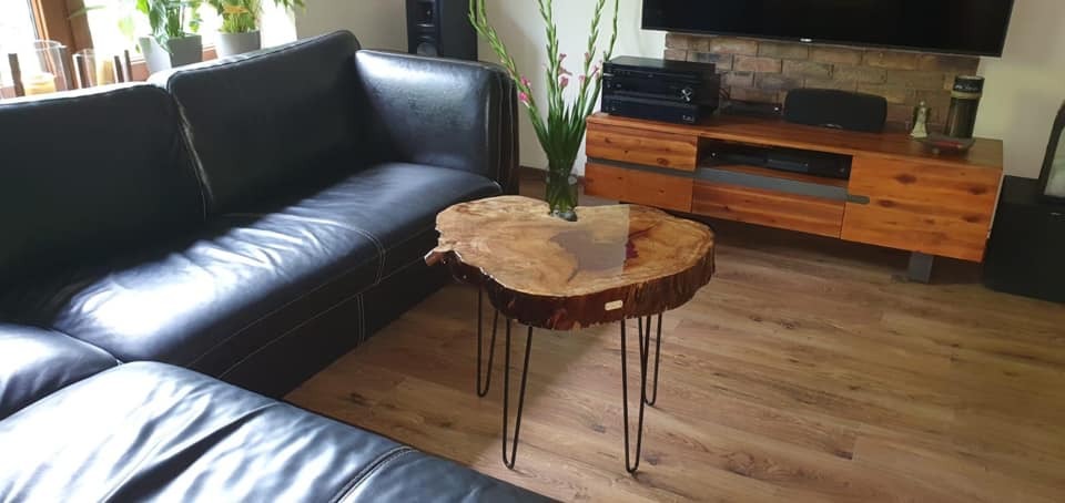 Stolik kawowy z blatem z naturalnego drewna z żywicą epoksydową, osadzony na trzech metalowych nogach typu hairpin, obok czarnej skórzanej sofy w salonie z drewnianą podłogą i szafką RTV z ceglaną...