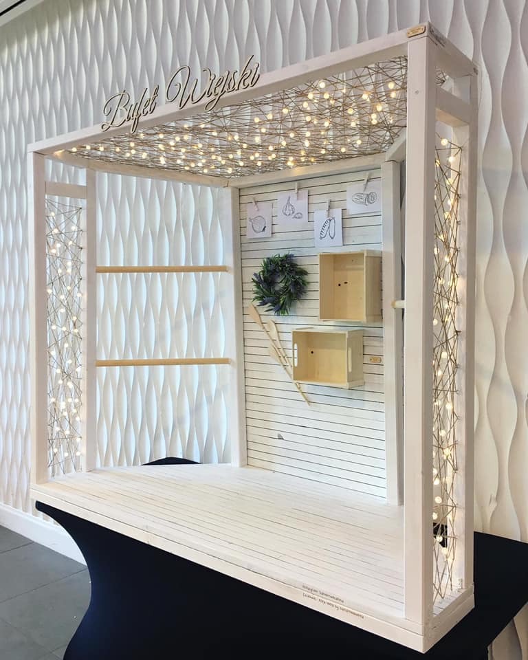 Drewniany bufet w stylu rustykalnym z napisem 'Bufet Wiejski', oświetlony girlandą żarówek, z dekoracjami w postaci wianka i rysunków warzyw.