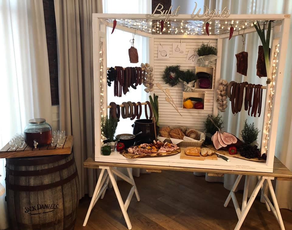 Dekoracyjny stół bufetowy z wędlinami, pieczywem i napojem, oświetlony lampkami, z napisem 'Bufet Wiejski' na górze, ustawiony na tle zasłon i beczki Jack Daniels.
