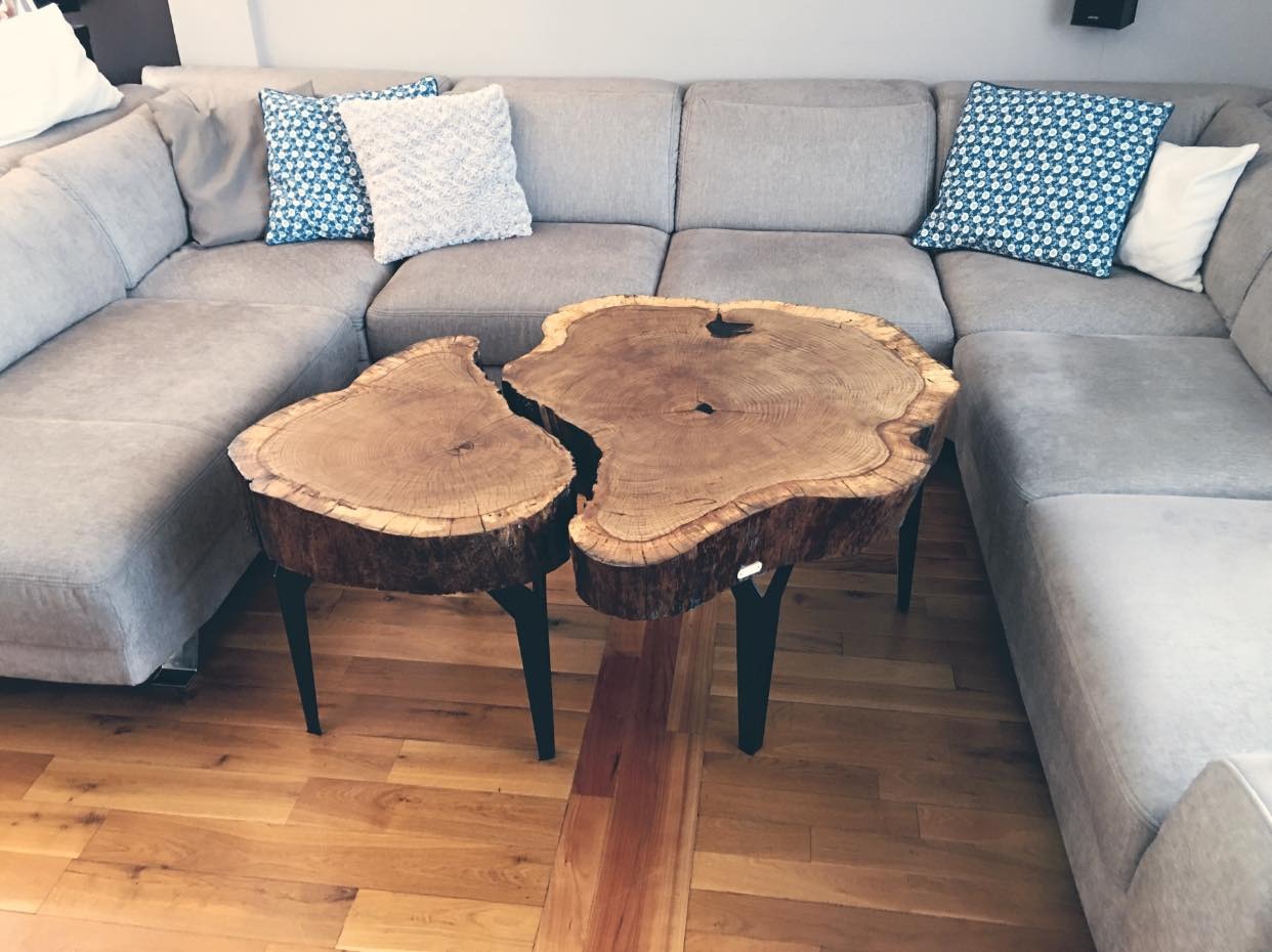 Stolik kawowy z dwóch plastrów drewna o nieregularnych kształtach, połączonych ze sobą, na czarnych, metalowych nogach, ustawiony przed szarą, narożną sofą z poduszkami w odcieniach błękitu i bieli...