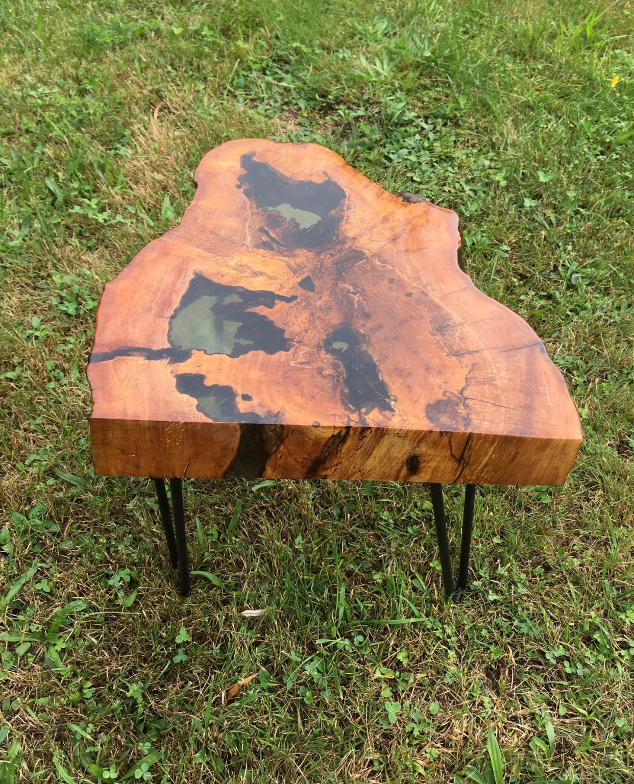 Niski stolik kawowy z blatem z litego drewna z żywicą epoksydową, na metalowych nóżkach, wykonany w Glinie, ustawiony na trawie.