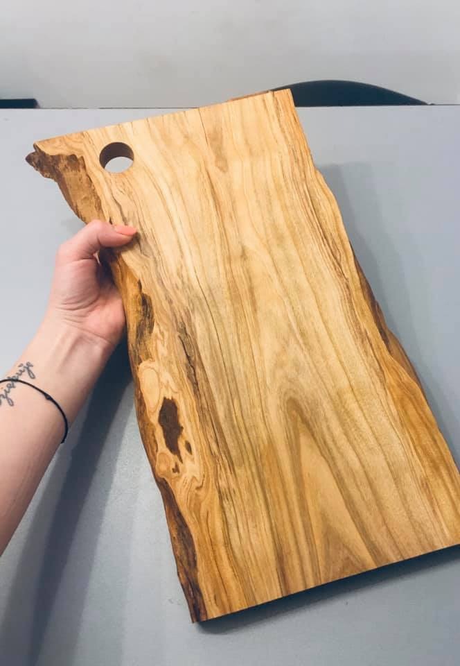 Deska do krojenia z jasnego drewna z naturalnymi krawędziami i wywierconym otworem, trzymana w dłoni z widocznym tatuażem na przedramieniu.