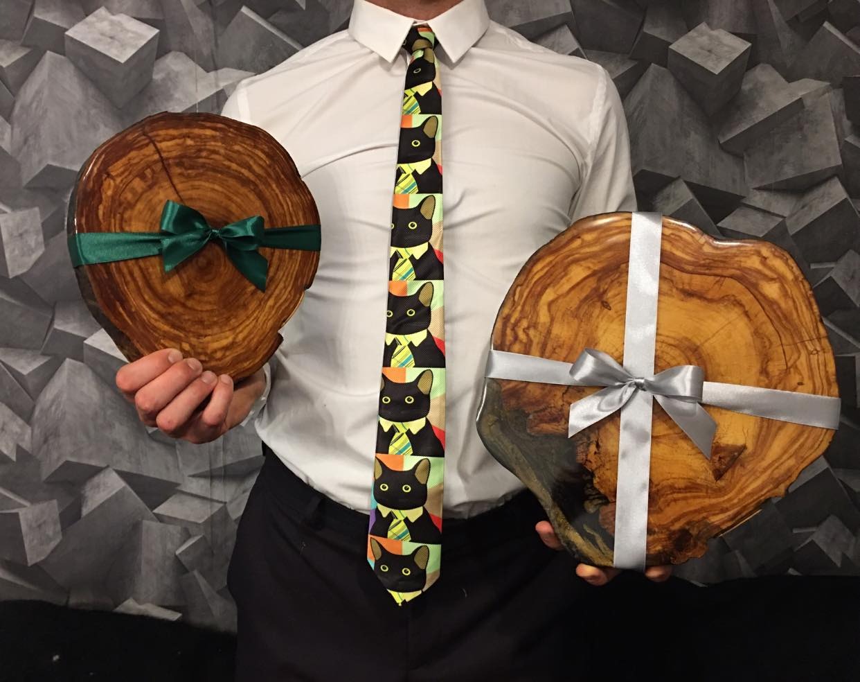 Mężczyzna w białej koszuli i krawacie w koty prezentuje dwa okrągłe, drewniane plastry z żywicą, przewiązane wstążkami, na tle ściany z geometrycznym wzorem.